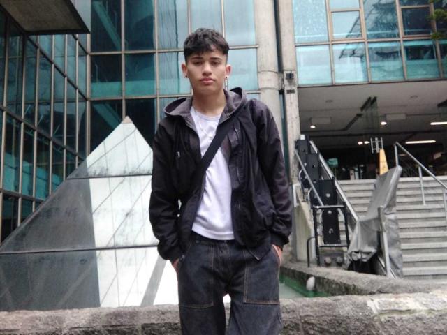 HarryClaude stands with hands in pockets, wearing a black jacket, white tee, and dark jeans. He has short dark hair and a neutral expression. Building with glass and metal.