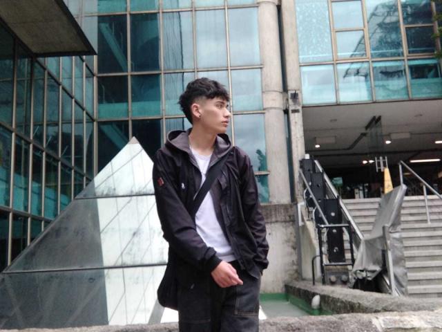 HarryClaude, standing with hands in pockets, wearing a black jacket and white shirt, looking to the side, in front of a glass building with stairs and metal structures.
