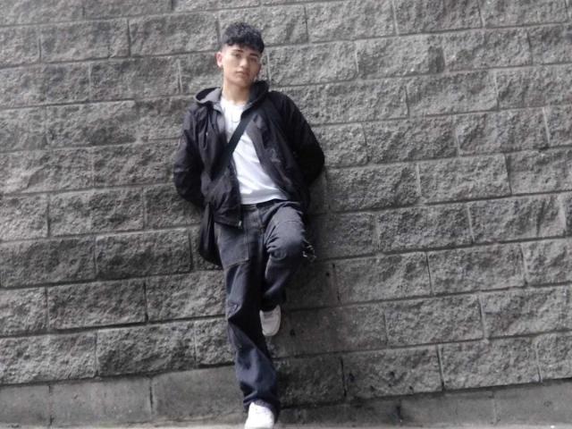 HarryClaude, standing against a grey brick wall, wearing a black jacket, white tee, and black pants, with hands in pockets, one leg raised, looking serious, black hair.