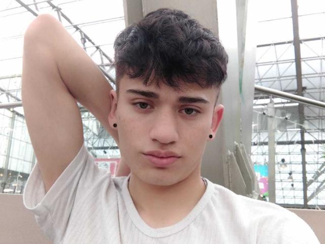 HarryClaude has curly black hair, small black ear studs, and a neutral expression. He's wearing a white tee, with his right arm behind his head, looking straight at the camera.