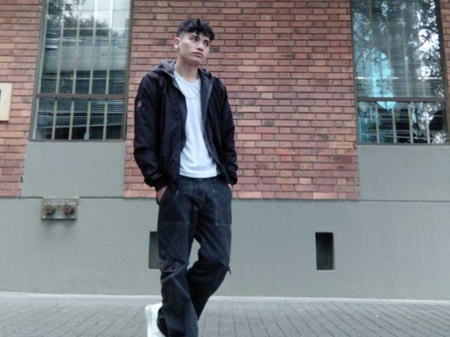HarryClaude is a skinny dude with dark hair, wearing a black jacket, white tee, and black pants, standing with hands in pockets, looking to the side.