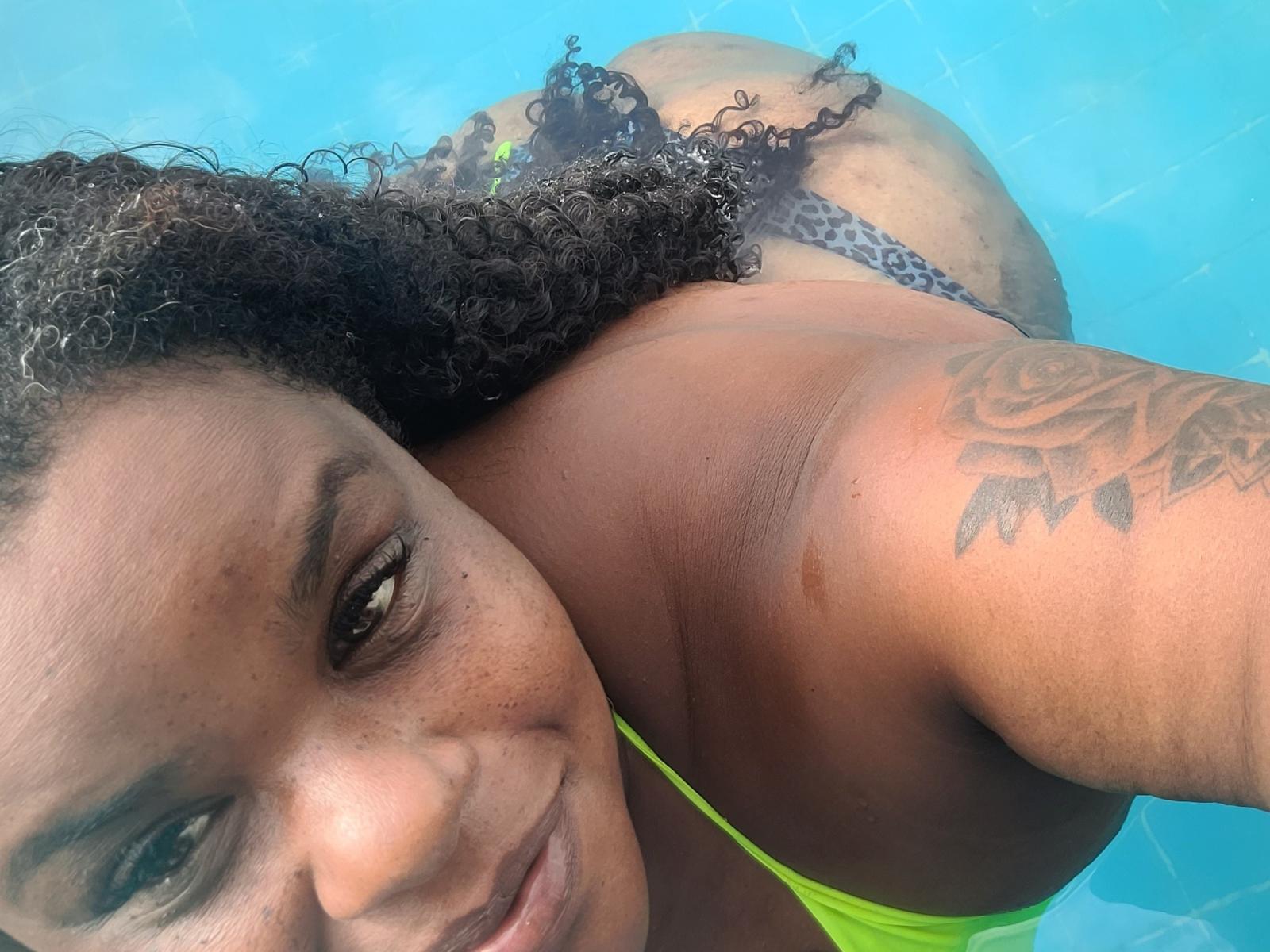 MonaRey in a pool, topless, with curly brown hair, green bikini bottom, and a rose tattoo on her right shoulder, looking at the camera with a slight smile.