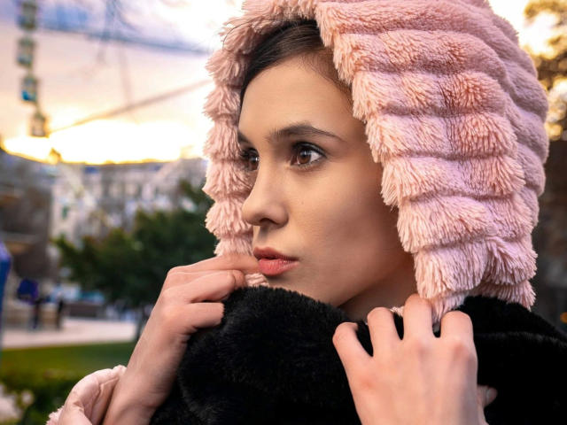 LegalKate-hot with pink, fluffy hood and black fur collar, hands near face, looking to the side. She has brown eyes, light skin, and a serious expression.