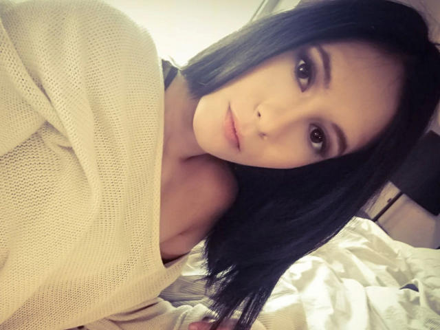 LegalKate - uma mulher atraente com cabelo preto, olhos castanhos e pele clara, vestindo um suéter de malha bege, olhando para a câmera com uma expressão ligeiramente séria, deitada em uma cama..