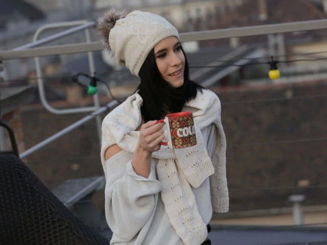 LegalKate-hot with dark hair, white knit hat, and oversized sweater, holding a red "COOL" mug, smiling on a rooftop with string lights in the background.