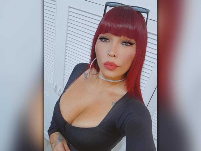 IrisGoddesss com cabelo curto vermelho, vestido preto, decote, tatuagens, colar e óculos de sol na cabeça, olhando para a câmera com os lábios curvados. Seios proeminentes no vestido. Fundo com porta branca com ripas..