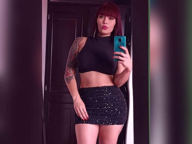 IrisGoddesss with red hair, black crop top, and sparkly black mini-skirt, tattoo on right arm, holding blue phone, standing in front of dark door, showing her ass.