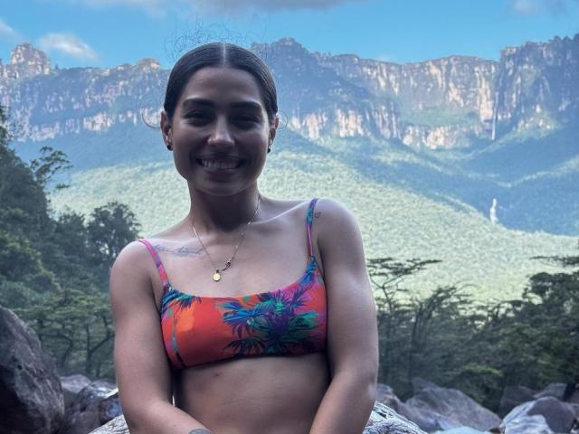 SamanthaBss with small tits, wearing a colorful pineapple-patterned bikini top, smiling, has tattoos on her left shoulder, and a gold necklace, standing in front of a mountain backdrop.