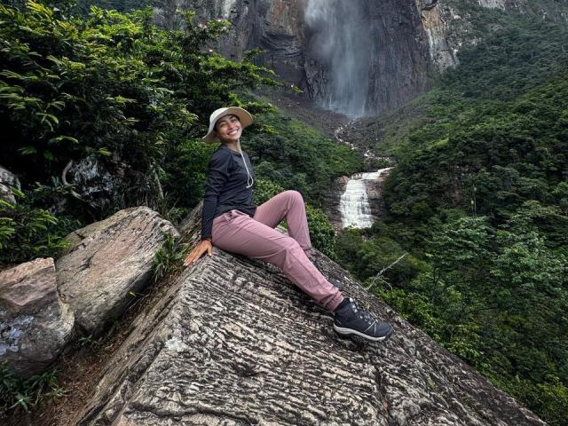 SamanthaBss with pink pants and black boots sits on a rocky ledge, smiling, wearing a hat, next to a waterfall, surrounded by green foliage. Her ass is slightly raised.