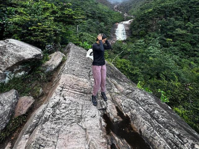 SamanthaBss with black shirt, pink pants, and black shoes stands on rocky cliff, hand in hair, green forest and waterfall in background, holding white backpack.