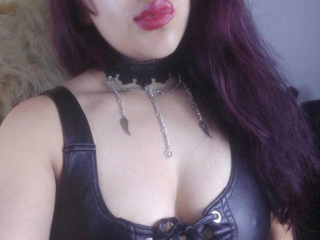 MariaMendozaI with dark purple hair, pouty red lips, black leather bra, and metal chain choker, showing her boobs. Her skin is light, and she's looking down.