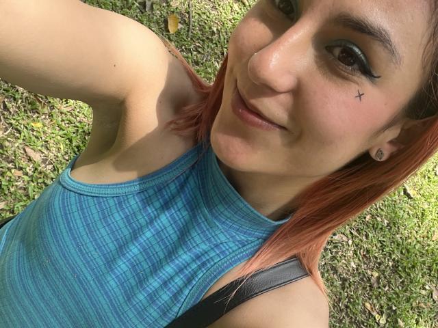 AmyStreetwise69 with pink hair, blue checkered sports top, black strap, small nose x, smiling, grassy background, armpit visible, brown eyes, black eyeliner, small earrings.
