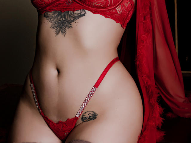EmmaRosseti's red lace bra and thong, showing her flat stomach, tattooed rose on her belly, skull tattoo on her left hip, and a sheer red robe.