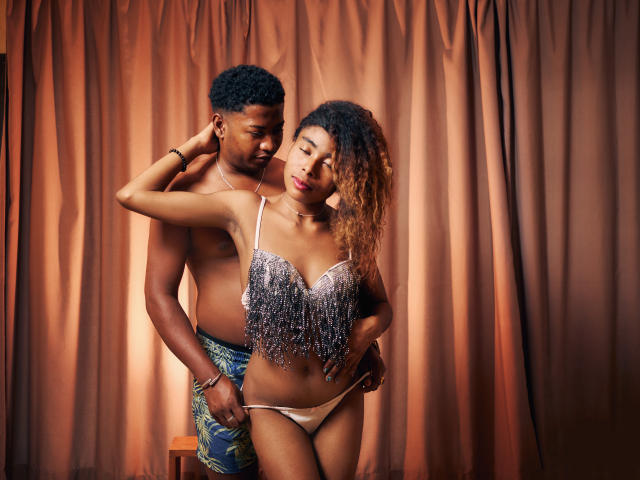 FredSyl with curly brown hair in a silver fringed top and white panties, standing in front of a shirtless black guy with green pants, his hand on her ass.
