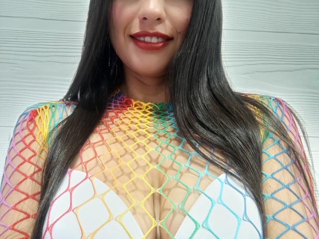 Madisson Fontaine, con cabello negro largo, lápiz labial rojo y una blusa de malla de colores como un arcoíris sobre su escote blanco, sonriendo. Sus pechos son visibles a través de la malla..