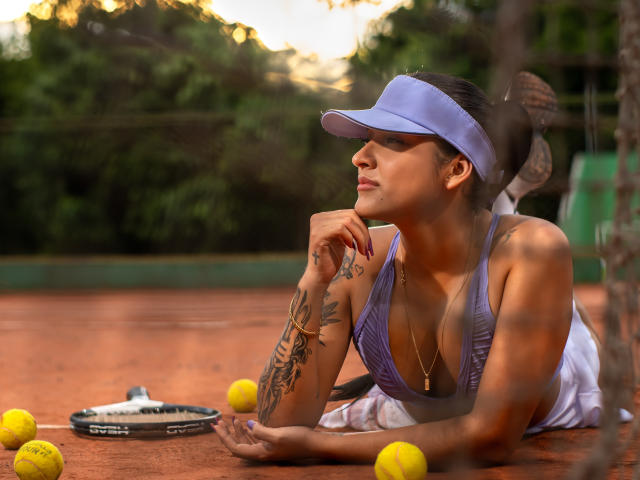 MiaSFoxy with tattoos, lying on a tennis court, wearing a purple visor and sports bra, showing her tits, surrounded by yellow tennis balls and a racket.