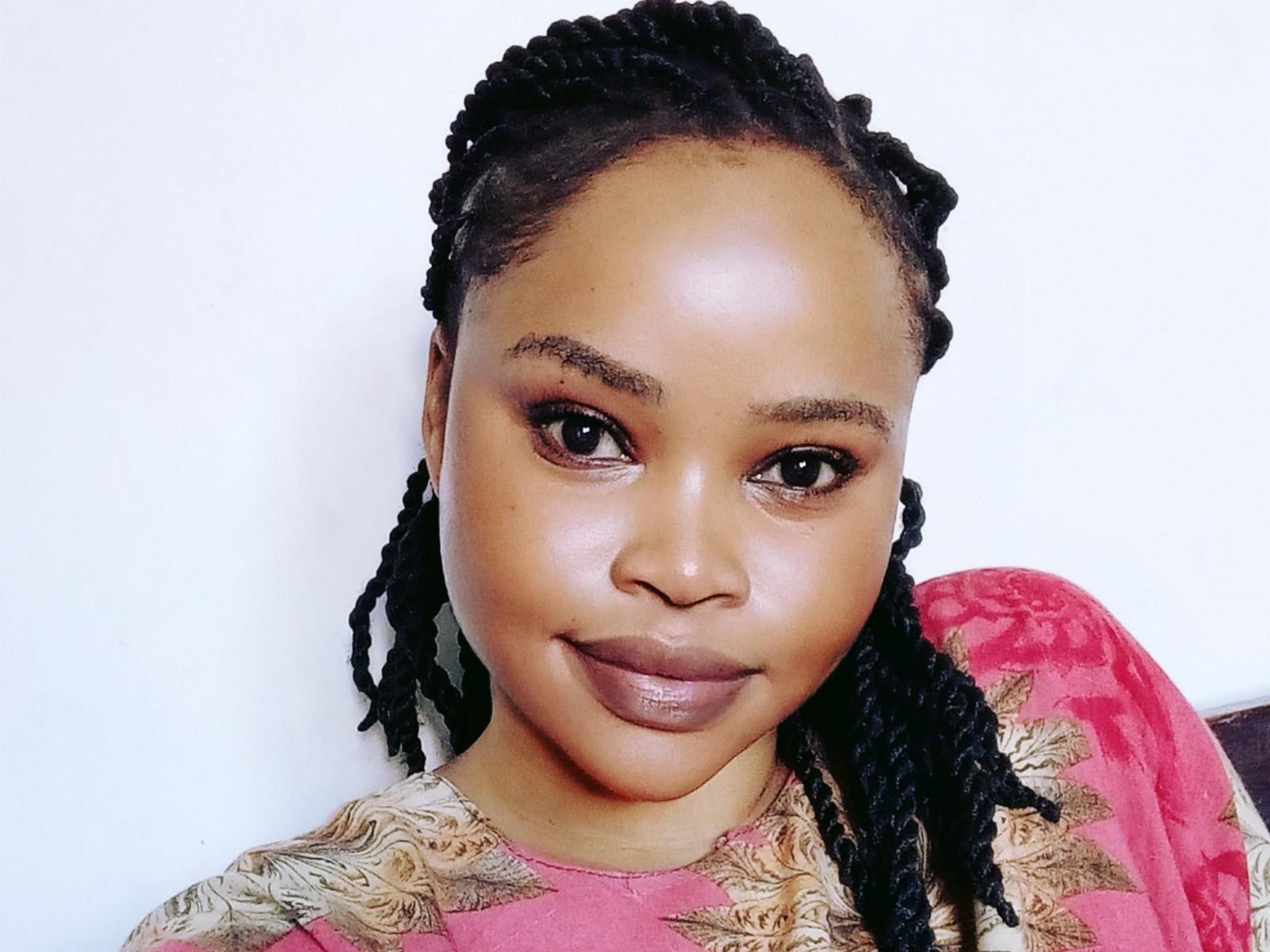 AfricanaQueen with dark brown skin, shiny black braided hair, full lips with brown lipstick, and brown eyes, wearing a red and gold patterned shirt.