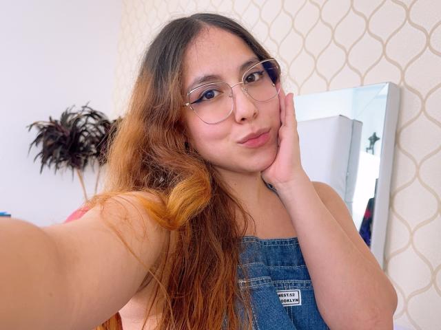 AmberlStha with long brown hair, glasses, and denim overalls, pouting with her hand on her cheek, showing her cute face and small tits. She's taking a selfie.