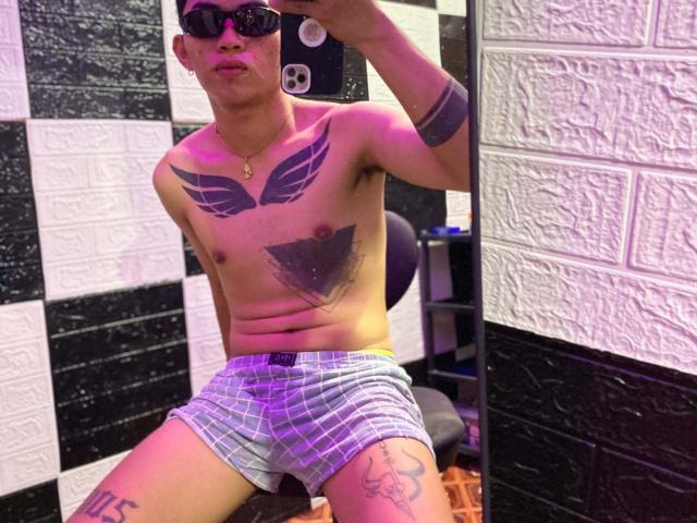 Shirtless CockOfAsian with tattoos, wearing plaid boxer shorts, black sunglasses, and a necklace, sitting in a room, holding a phone, with a relaxed, confident expression.
