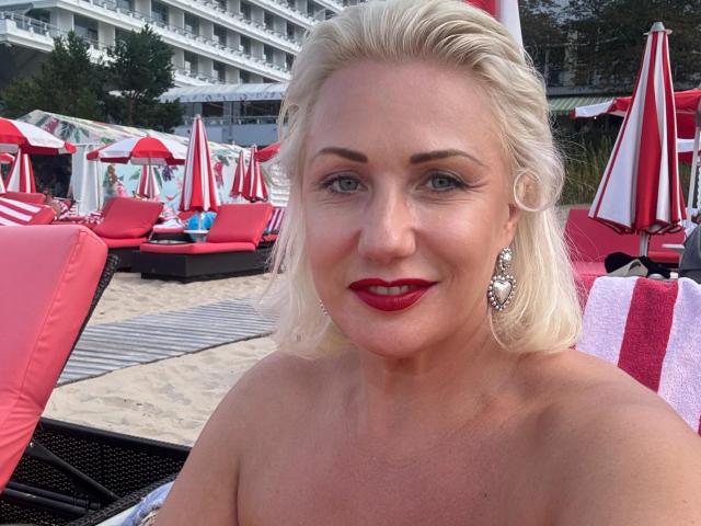 Cheryl, con cabello rubio, lápiz labial rojo y aretes rojos, está sentada sobre una toalla roja en una playa con sombrillas rojas y blancas, mostrando sus hombros desnudos..