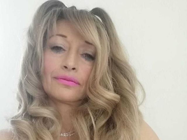 BigBoobsy69 has blonde curly hair in pigtails, pink lipstick, and a subtle smile. She's wearing a silver necklace against a plain white background.