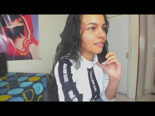 CanelitaRousse with wet black hair, talking on a phone, wearing a white and black "DREAM" hoodie, smiling slightly, in a bedroom with colorful bedspread.