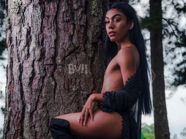 KattaleyaHorny with dark long hair, tan skin, and no clothes except black lace sleeves and thigh-high boots, leaning against a tree with a serious expression, showing her ass.