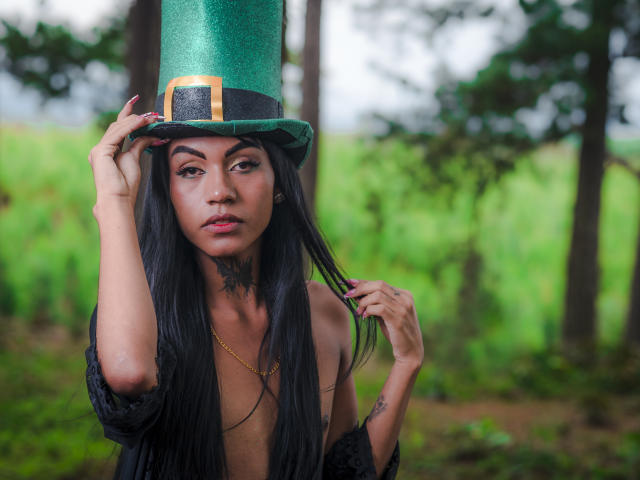 KattaleyaHorny wearing a green top hat with a gold buckle, long black hair, tattoos, and a gold necklace, touching her hat, in a forest, with a serious expression.