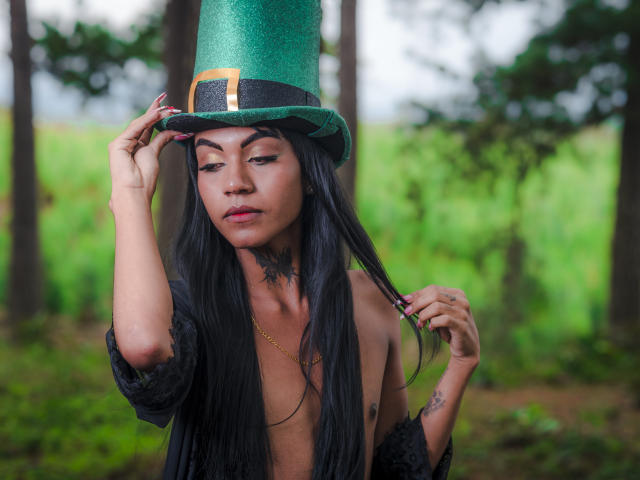 KattaleyaHorny with long black hair, wearing a green glitter top hat and black lace shawl, touching her hat, showing tattoos and a gold necklace, in a forest.