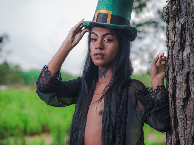 KattaleyaHorny with long black hair, green top hat, black lace robe, and visible nipples, standing by a tree, touching hat with left hand, right hand on tree.