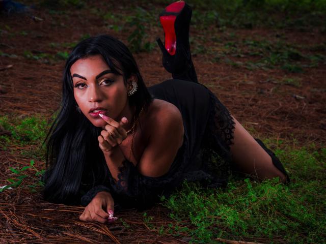 KattaleyaHorny, with long black hair, wearing a black lace dress and red-soled high heels, lying on pine-covered ground, sucking her finger, with her ass up and looking at camera.
