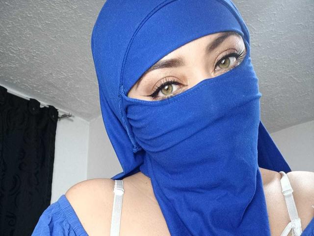 Zahra Jalid, mit blauem Kopftuch und Maske, wobei ihre grünen Augen und weiße BH-Träger sichtbar sind, hat helle Haut und dunkle Augenbrauen. Das Foto zeigt nur ihr Gesicht und ihre oberen Schultern..