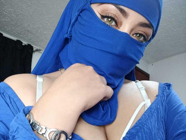 ZahraJalid with green eyes, wearing a blue hijab and mask, exposing white bra straps and cleavage, holding blue cloth over mouth, silver bracelet on right wrist.