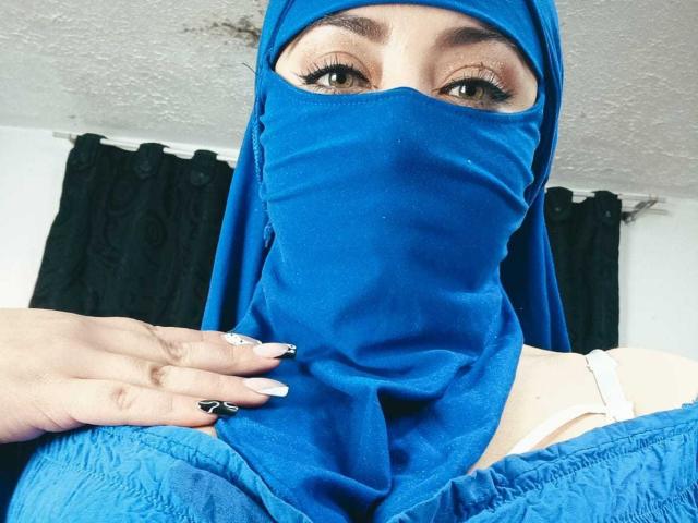 ZahraJalid with blue hijab and matching mask, exposing brown eyes. She's holding her mask, showing white nails with black and white designs, and beige bra.