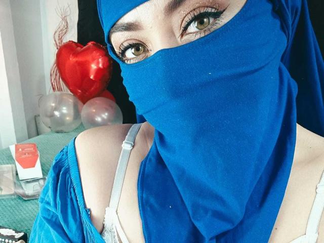ZahraJalid with blue hijab covering her mouth and nose, green eyes looking at the camera, white bra straps visible, red heart balloon in the background.
