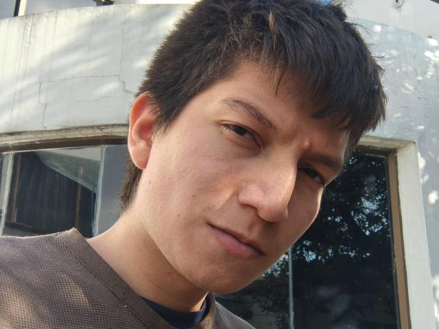 ErossGuy has a slightly dirty face, short brown hair, and a small nose with a subtle smirk. Wearing a brownish shirt, standing in front of a window.