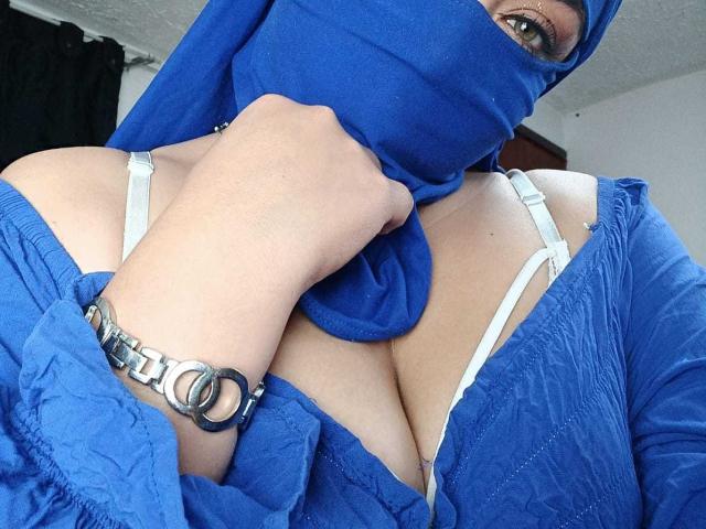 ZahraJalid with blue mask, blue dress, and white bra showing her big tits, holding dress up, wearing a chain bracelet with metal rings, green eyes visible.