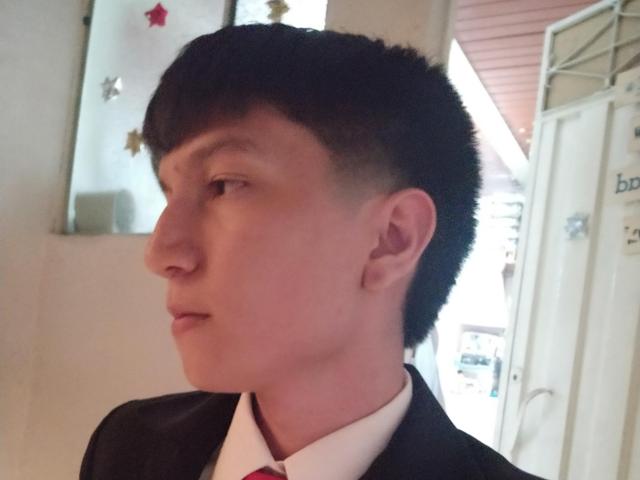 ErossGuy with a short, dark, buzzcut hairstyle, pale skin, and a serious expression, wearing a black suit jacket and white shirt with a red tie.