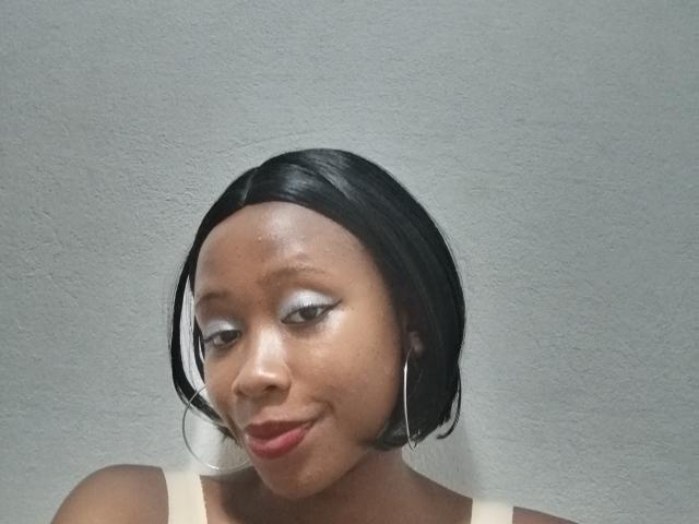 HoneyT with dark skin, black bob, white eyeliner, red lipstick, and silver hoop earrings, smiling slightly, wearing a beige top, in a simple, textured gray background.