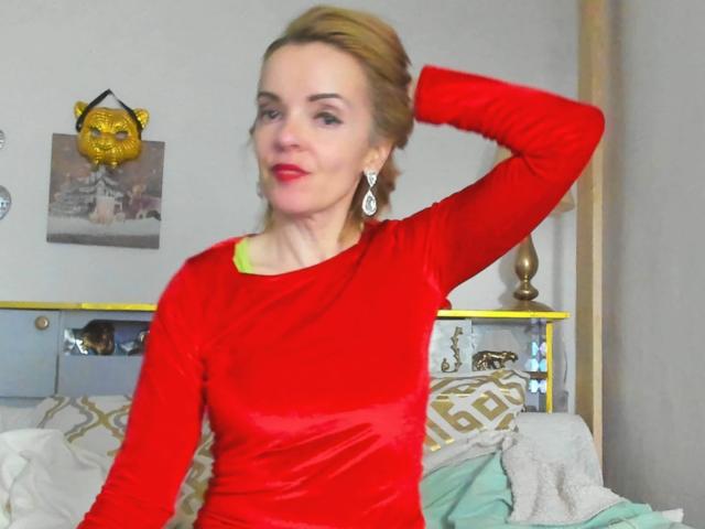 KateRouge has red velvet long-sleeve shirt, red lipstick, earrings, blonde hair in bun, hand behind head, sitting on bed with white and green pillows.