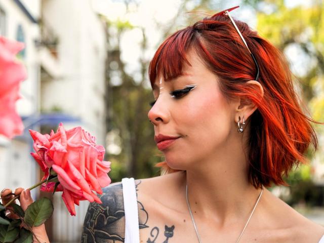 AriiannaHot with red hair, eyeliner, and tattoos, holding a pink rose, looking down at it, wearing a white strap, silver necklace, and red sunglasses on her head.