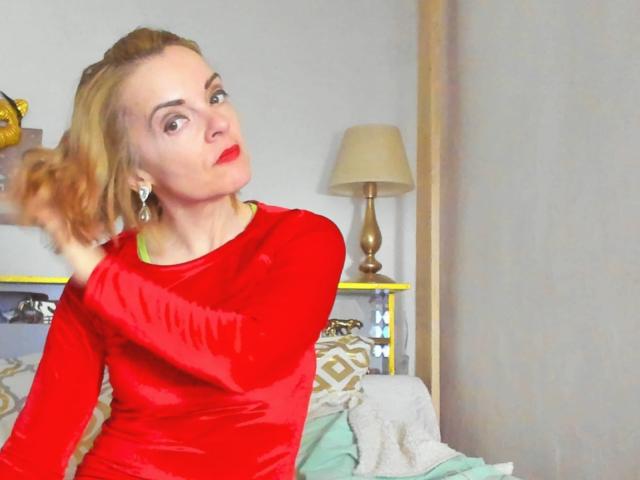KateRouge with blonde hair, red velvet long-sleeve dress, and red lipstick, holding her hair, looking surprised, sitting on a bed with green and white pillows.