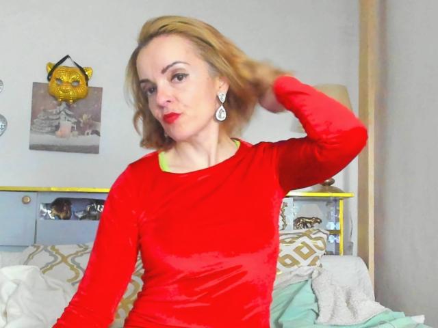 KateRouge in a shiny red velvet top, holding her blonde hair with one hand, wearing green earrings and red lipstick, sitting on a bed with yellow and white pillows.