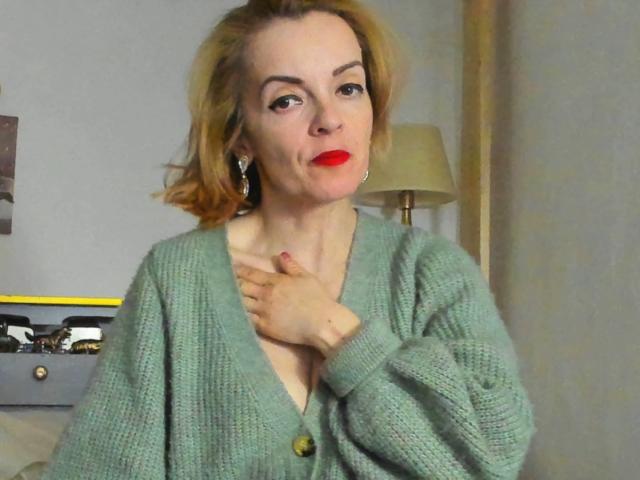 KateRouge has red lipstick, green cardigan, and short blonde hair, with hand on chest, looking slightly pouty. She's wearing earrings, and has a fair skin tone.