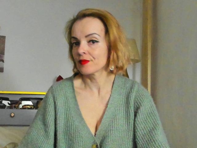 KateRouge has red lipstick, green knit cardigan, and short blonde hair. She's smiling with visible cleavage, wearing pearl earrings. Background is blurry. Image is slightly blurry too.