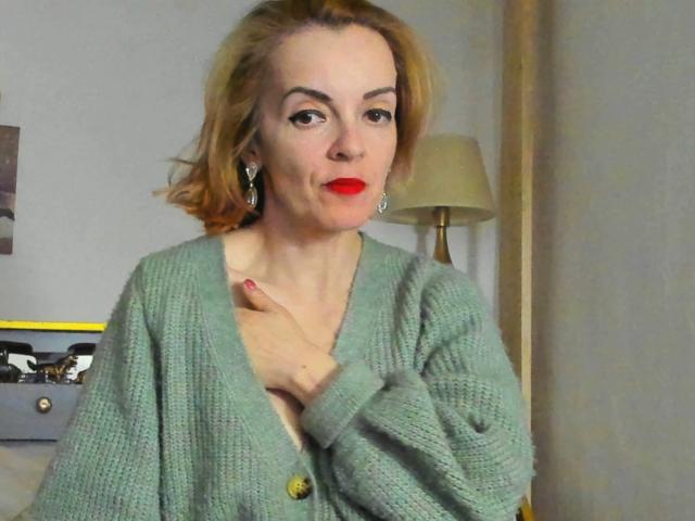 KateRouge has red lipstick, short blonde hair, and wears a green knitted cardigan. She's touching her chest with her right hand, looking slightly pouty.