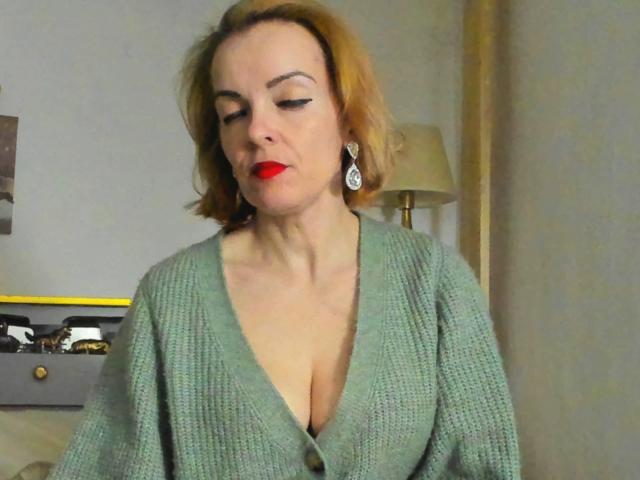 KateRouge with red lipstick, green knit cardigan, deep cleavage, and teardrop earrings, looking down with a slightly pensive expression. Her short brown hair is slightly messy.