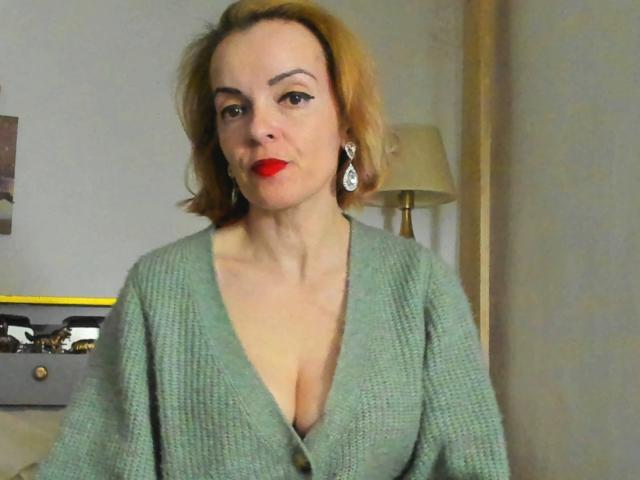 KateRouge with red lipstick, green knitted sweater, showing her tits, short brown hair, earrings, sitting, looking at camera, lamp in background, light-colored wall, relaxed expression.