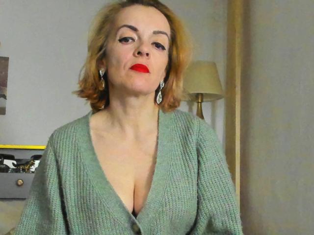 KateRouge has red lipstick, short blonde hair, and wears a green knit sweater with a deep V-neck, showing her boobs. She's wearing earrings and looking slightly up.