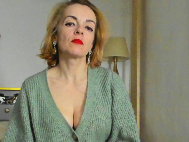 KateRouge with short blonde hair, red lipstick, and green knitted sweater, showing her cleavage, wearing dangling earrings, sitting in a room with a lamp behind her.